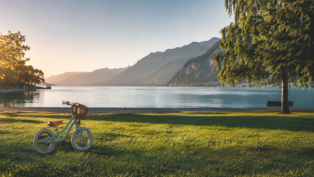 Petit vélo d'enfant posé dans l'herbe au bord du lac d'Annecy au lever du jour, montagnes en arrière-plan