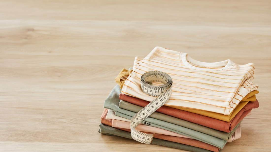 Pile de t-shirts enfant pliés aux tons écru, terracotta et vert sauge avec un mètre ruban de couturière