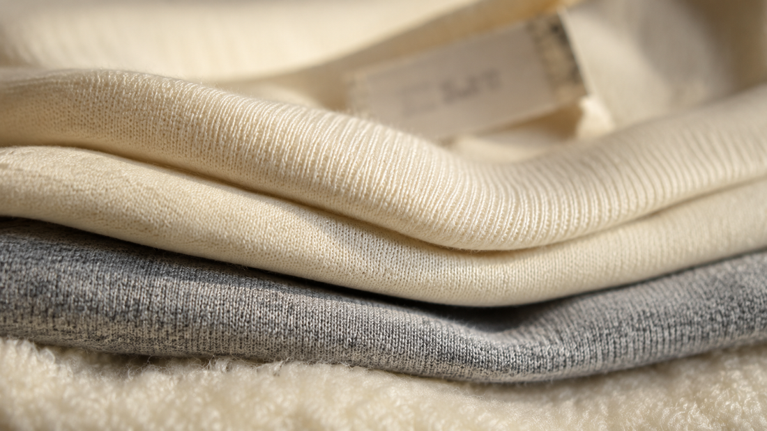 Gros plan sur trois textures de tissu superposées, jersey écru, gris chiné et molleton gratté, avec une étiquette cousue