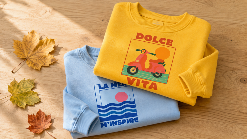 Deux sweats enfant pliés sur une table en bois, l'un jaune Dolce Vita, l'autre bleu ciel La Mer M'inspire